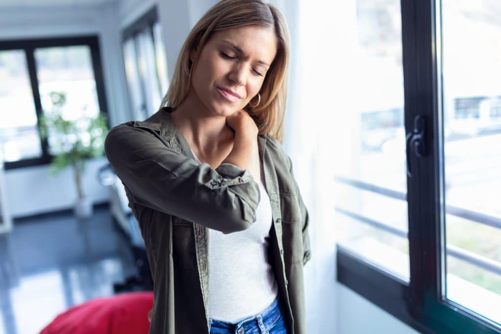 Shot of tired young woman with neck and back pain standing in the living room at home.