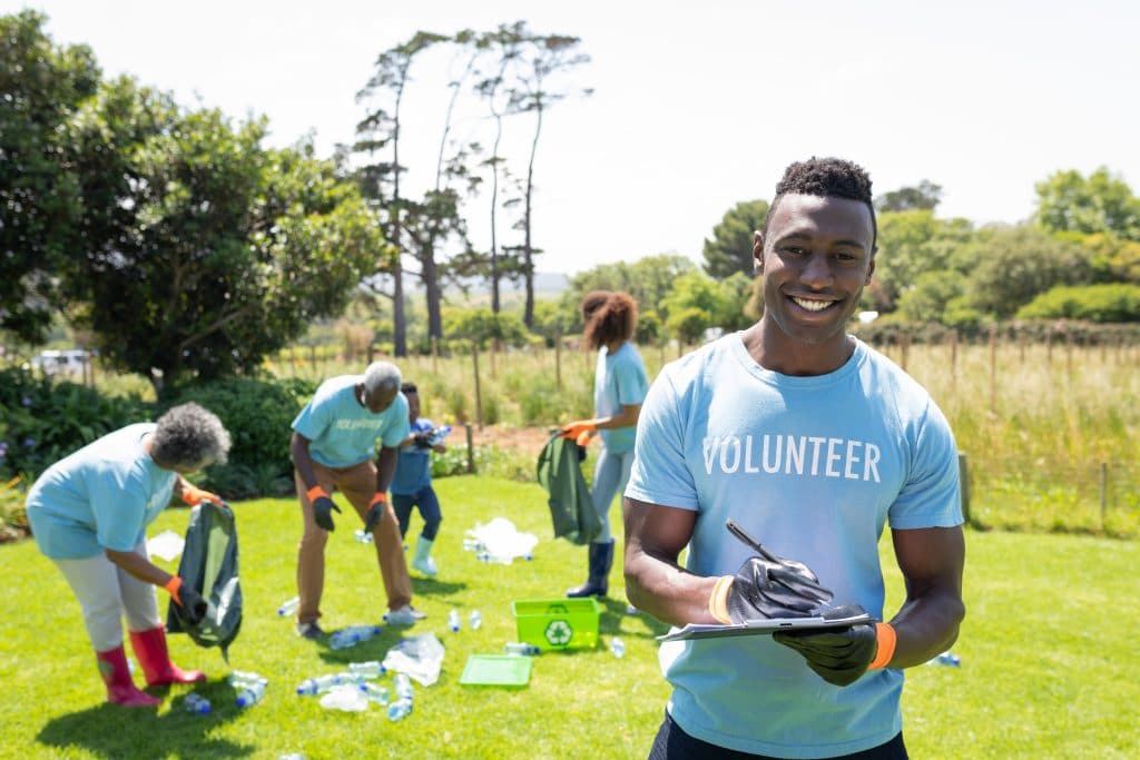 Portrait of a smiling young African American man standing in a field wearing wearing a t shirt with volunteer written on it and gloves, writing on a clipboard and smiling to camera, while a diverse group of volunteers collect rubbish and recycling in the background